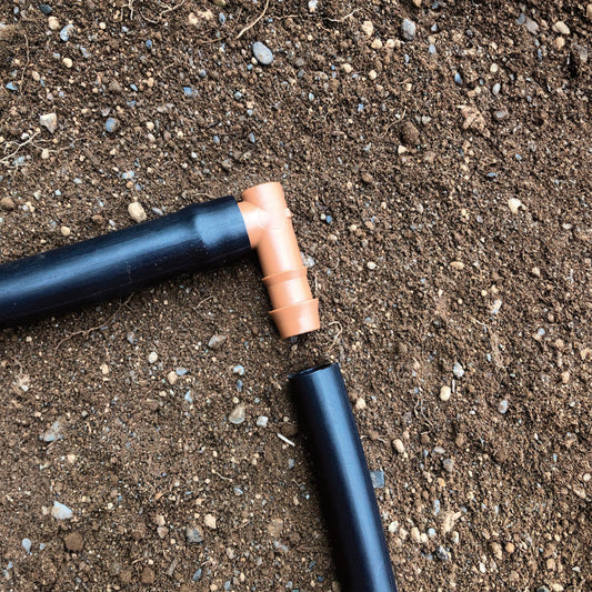 A brown 1/2" barbed drip tube elbow fitting connected to 1/2" drip tubing with soil in the background.