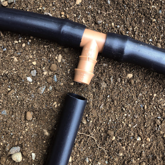 A brown 1/2" barbed drip tube tee fitting connected to 1/2" drip tubing with soil in the background.