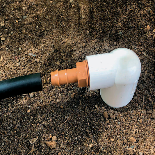 A brown barbed drip tube adapter with a 3/4-inch threaded connection connected to 1/2" drip tubing with soil in the background.