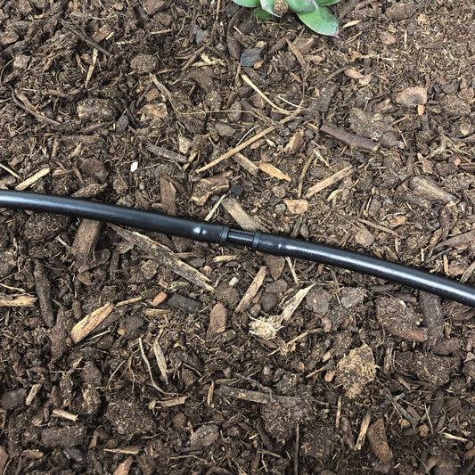 A black 1/4" barbed drip tubing coupler made of uv resistant plastic, used for repairing or connecting drip irrigation tubing. Coupler is connected with 1/4" drip tubing with soil and plants in the background.