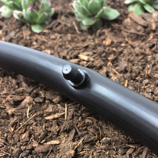 A black 1/4" barbed drip tubing plug made of uv resistant plastic, used for repairing drip irrigation tubing. Plug is connected to 1/2" drip tubing with soil and plants in the background.