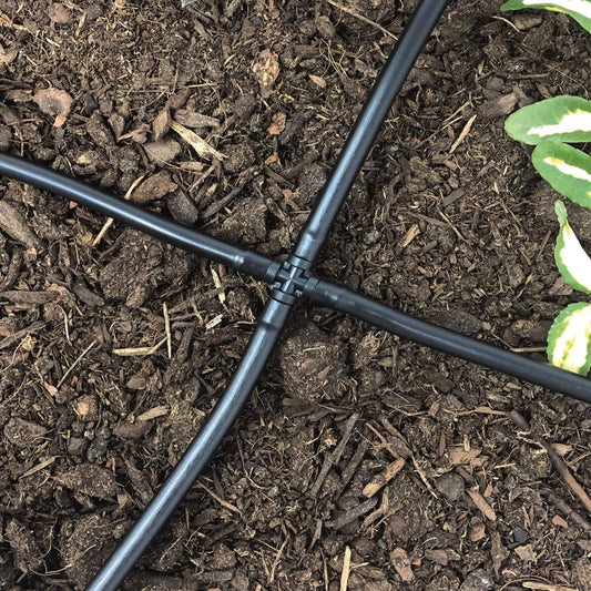 A black 1/4" barbed drip tubing cross cross made of uv resistant plastic, used for routing dripper lines in multiple directions with soil and plants in the background.
