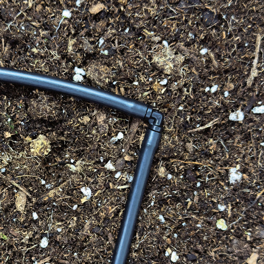 A black 1/4" barbed drip tubing elbow made of uv resistant plastic, used for routing drip irrigation. connected with 1/4" drip tubing with soil and plants in the background.
