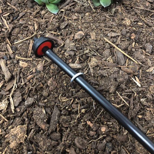 A 4-inch wire U-stake designed for 1/4-inch drip tubing with soil and plants in the background. Stake is holding 1/4" drip tube in place with drip emitter attached.