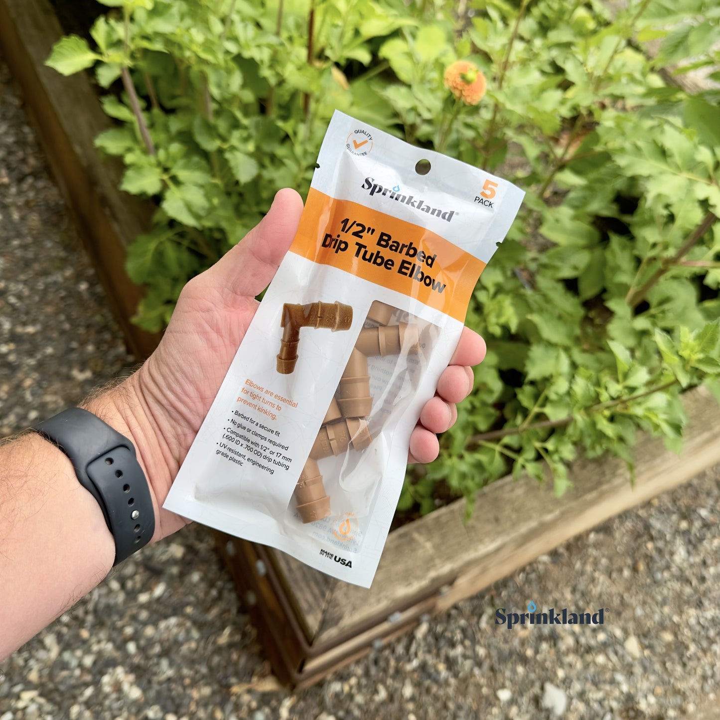 A 5 pack of brown 1/2" barbed drip tube elbow fitting with soil and plants in the background.