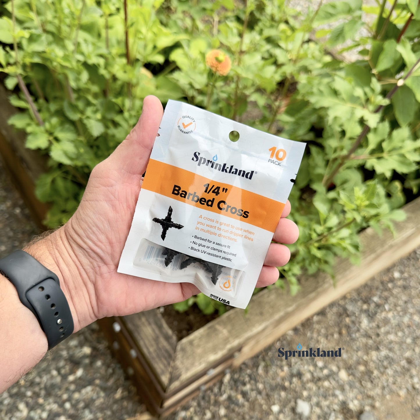 A 10 pack of black 1/4" barbed drip tubing cross cross made of uv resistant plastic, used for routing dripper lines in multiple directions with soil and plants in the background.