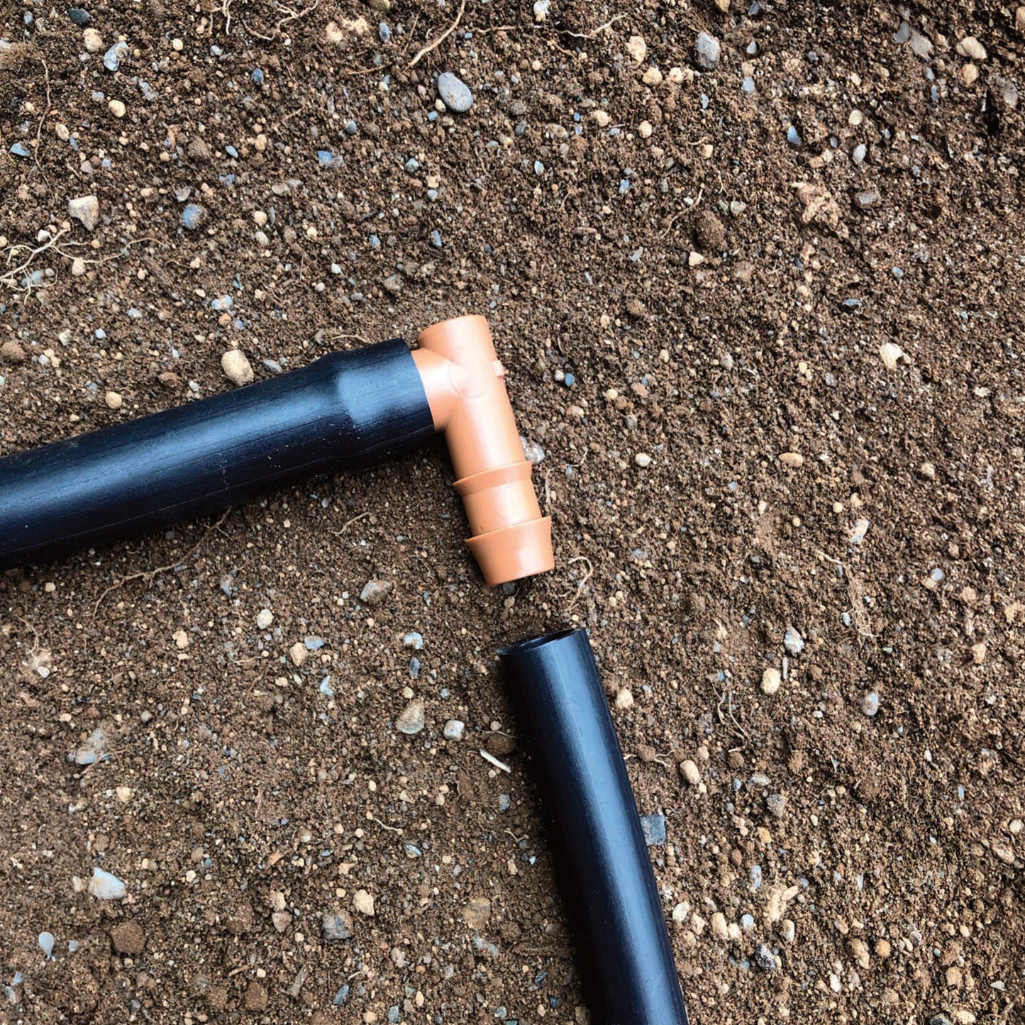 A brown 1/2" barbed drip tube elbow fitting connected to 1/2" drip tubing with soil in the background. 