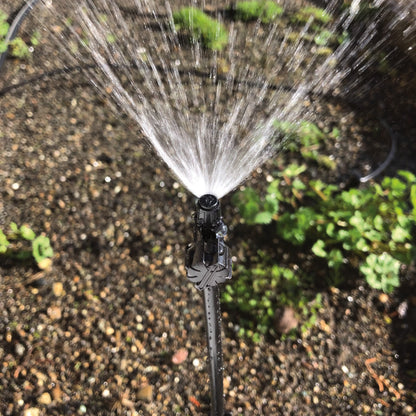 A black 90-degree spray head with adjustable flow, designed for use in lawn and garden irrigation systems with plants an soil in the background. 