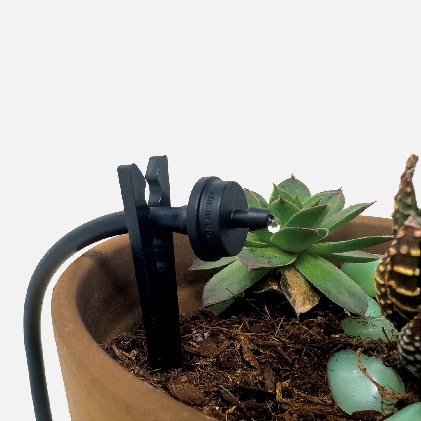 A single black 6" drip tubing plastic support stake designed to hold tubing in place with plants and soil in the background. 