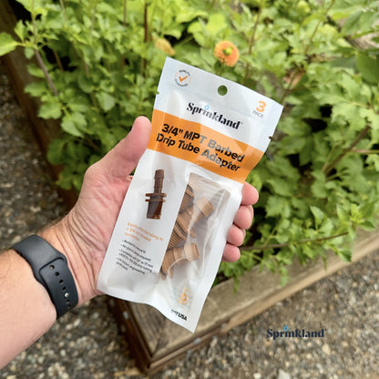 A 3 pack of brown barbed drip tube adapter with a 3/4-inch threaded connection connected to 1/2" drip tubing with soil and plants in the background.