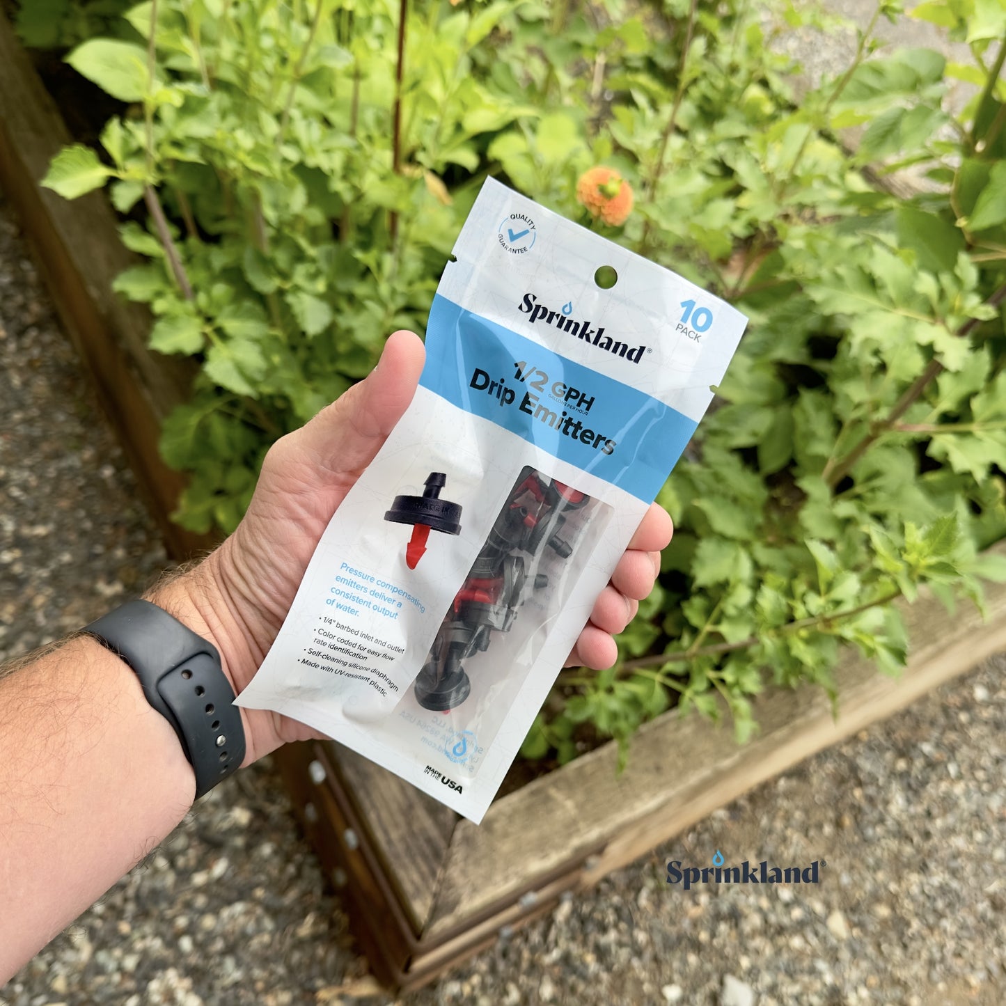 A 10 pack of black and red, plastic 1/2 GPH drip emitter for sprinklers with a 1/4" barb inlet and outlet, color-coded for flow rate identification with plants and soil in the background.