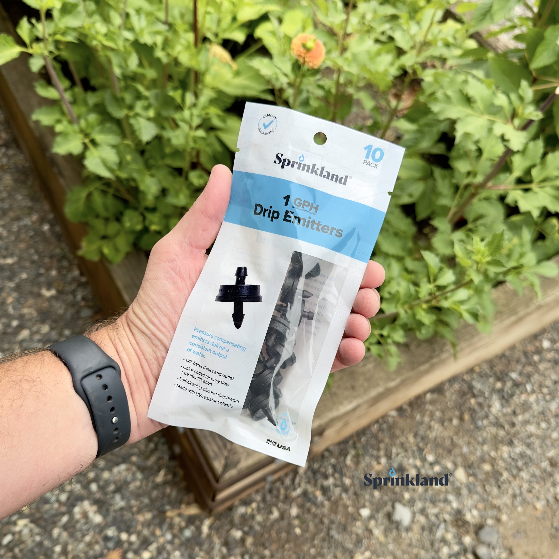 A 10 pack of black, plastic 1 GPH drip emitters for sprinklers with a 1/4" barb inlet and outlet, color-coded for flow rate identification with plants and soil in the background.