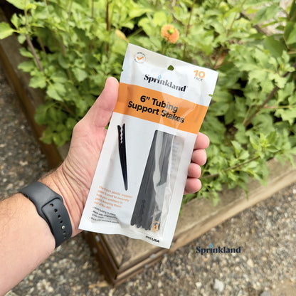A 10 pack of  black 6" drip tubing plastic support stake designed to hold tubing in place with plants and soil in the background. 